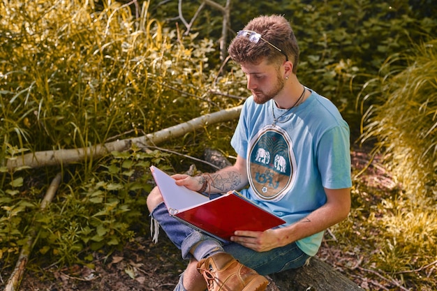 Blonde student die zijn aantekeningen bekijkt met een notitieboekje zittend op een houtblok midden in de natuur