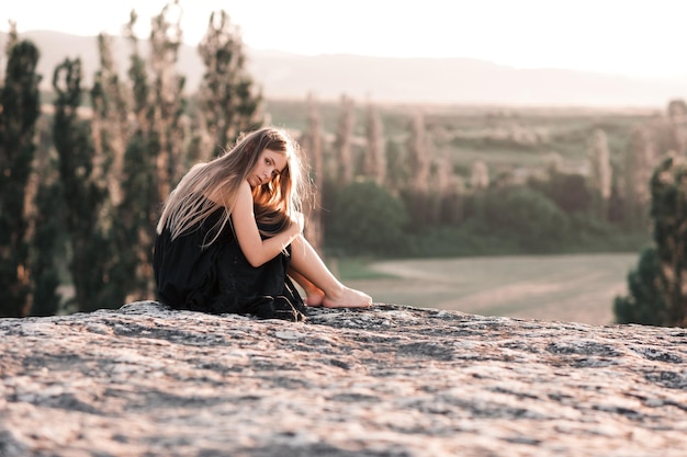 Blonde teen girl sitting alone on rock over beautiful nature background