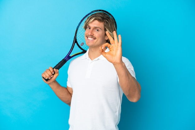 손가락으로 확인 표시를 보여주는 파란색 배경에 고립 된 금발 테니스 선수 남자