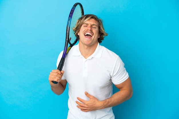 Blonde tennis player man isolated on blue wall smiling a lot