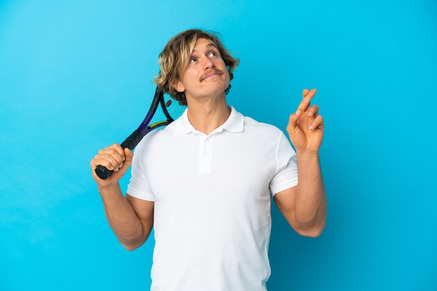 Blonde tennis player man isolated on blue with fingers crossing and wishing the best