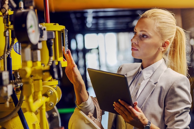 Blonde toegewijde succesvolle zakenvrouw in formele slijtage die machines controleert en tablet vasthoudt terwijl hij in een elektriciteitscentrale staat