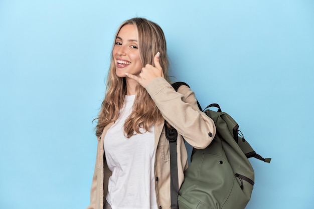 Blonde traveler with a travel backpack showing a mobile phone call gesture with fingers