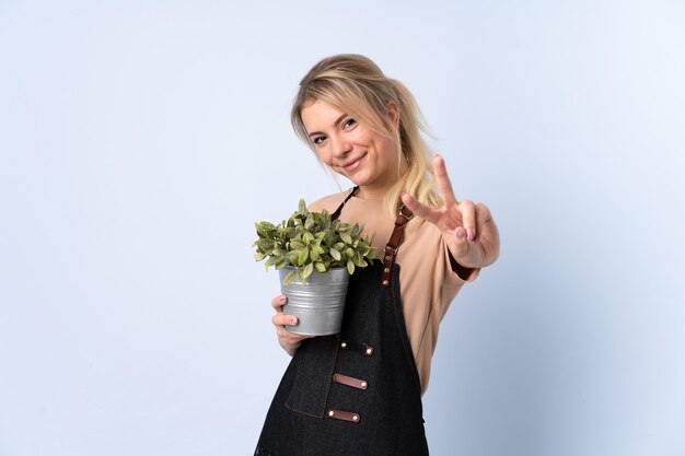 Blonde tuinmanvrouw die een plant over geïsoleerde muur houdt die en overwinningsteken glimlacht toont