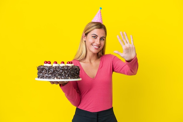 Blonde Uruguayan girl holding birthday cake isolated on yellow background counting five with fingers