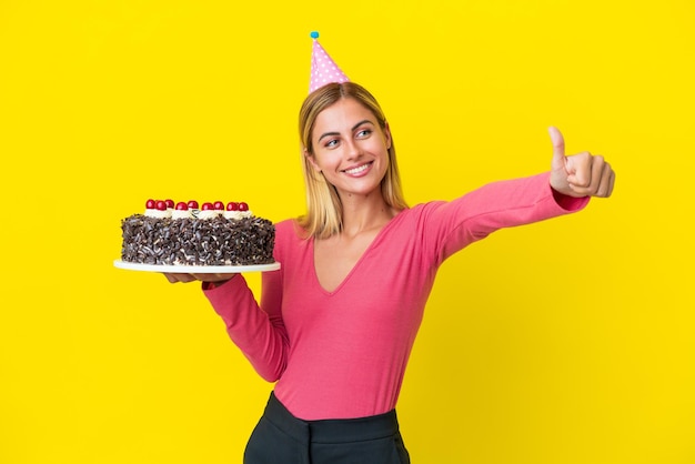 親指を立てるジェスチャーを与える黄色の背景に分離されたバースデーケーキを保持しているブロンドのウルグアイの女の子