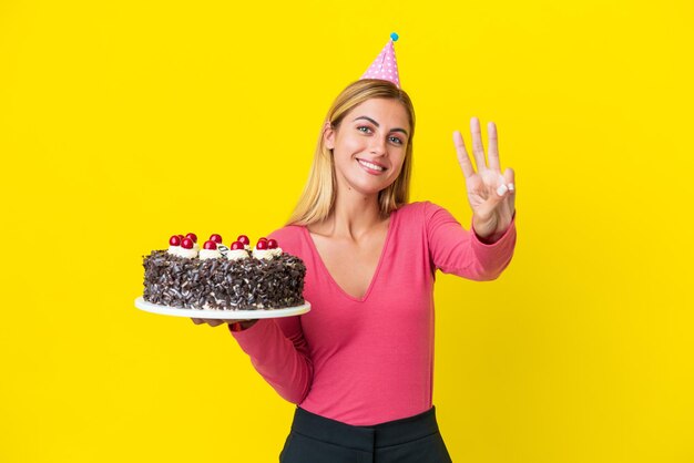 幸せな黄色の背景で隔離の誕生日ケーキを保持し、指で3を数えるブロンドのウルグアイの女の子