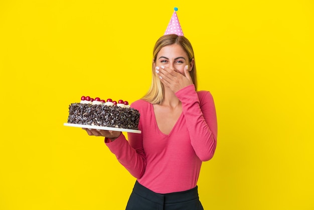 Блондинка из Уругвая держит торт ко дню рождения на желтом фоне счастливая и улыбающаяся, прикрывая рот рукой