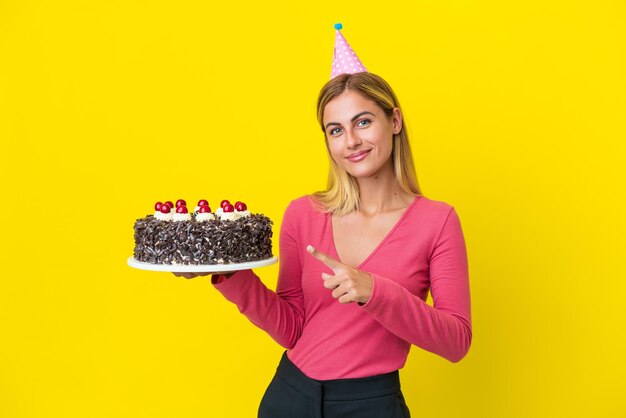 Ragazza bionda uruguaiana con torta di compleanno isolata su sfondo giallo che indica il lato per presentare un prodotto