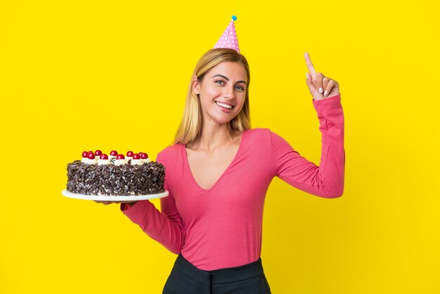 Blonde Uruguayan girl holding birthday cake isolated on yellow background pointing up a great idea