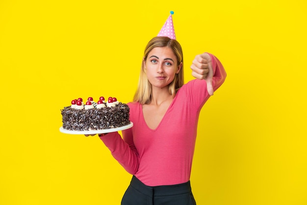 Blonde Uruguayan girl holding birthday cake isolated on yellow background showing thumb down with negative expression