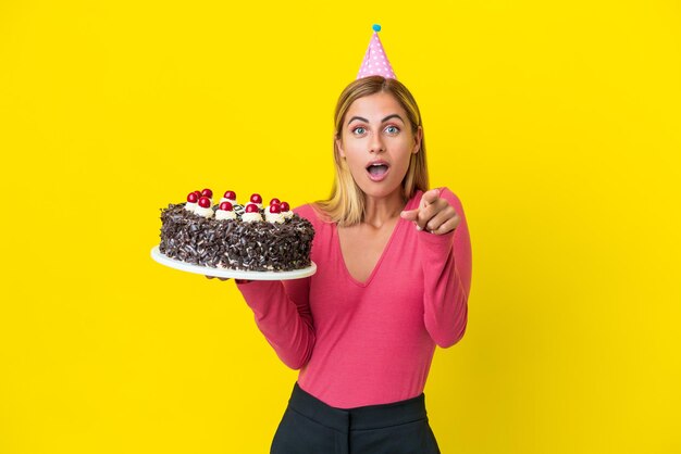 Blonde uruguayan girl holding birthday cake isolated on yellow background surprised and pointing front