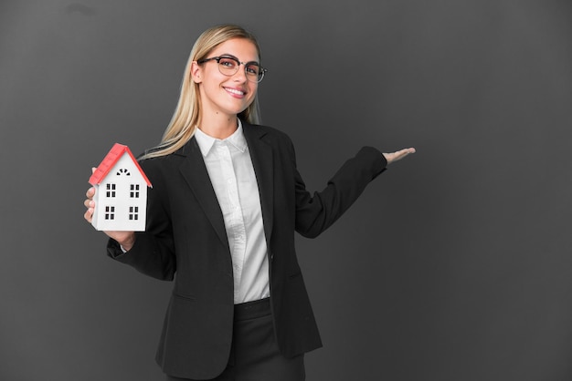 Blonde Uruguayan girl holding a house toy isolated on black background extending hands to the side for inviting to come