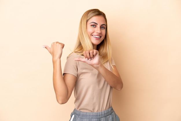 Blonde Uruguayan girl isolated on beige background pointing to the side to present a product