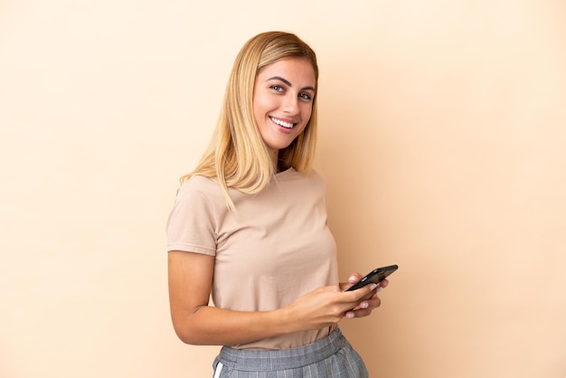 Blonde Uruguayan girl isolated on beige background sending a message with the mobile