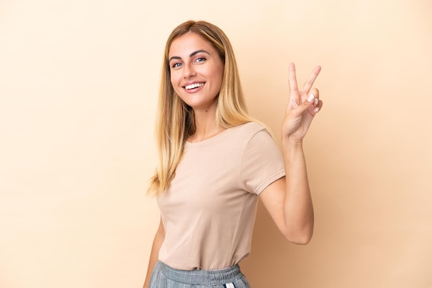 Blonde Uruguayan girl isolated on beige background smiling and showing victory sign