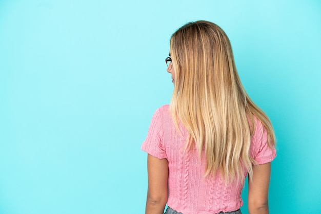 Blonde Uruguayan girl isolated on blue background in back position and looking side