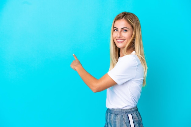 Blonde Uruguayan girl isolated on blue background pointing back