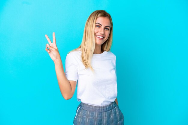 Blonde Uruguayan girl isolated on blue background smiling and showing victory sign