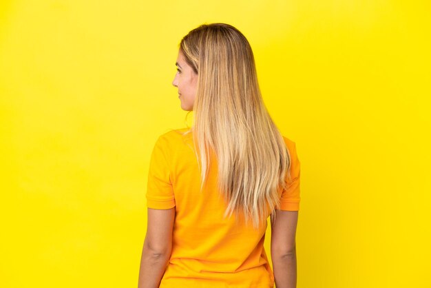 Blonde Uruguayan girl isolated on yellow background in back position and looking side