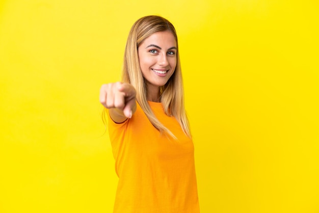 Blonde Uruguayan girl isolated on yellow background pointing front with happy expression