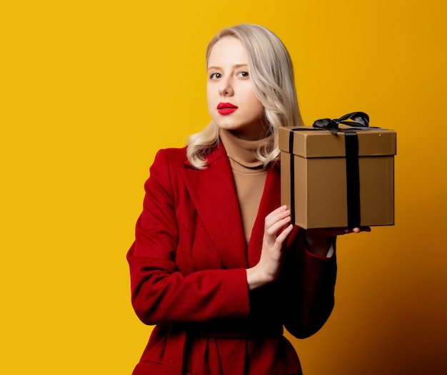 Blonde verraste vrouw in rode jas met giftdoos op gele muur