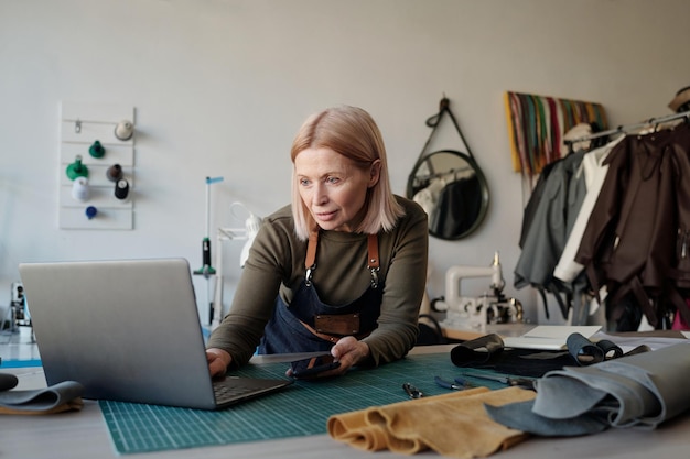 Blonde volwassen eigenaar van een leerbewerkingswinkel die naar het laptopscherm kijkt