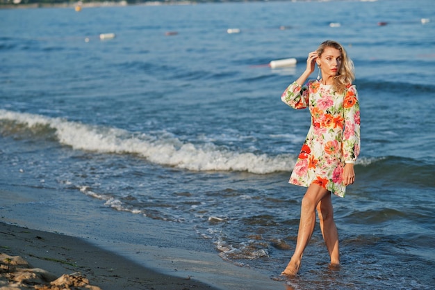 Blonde vrouw die een jurk draagt die op het zandstrand aan de kust staat en geniet van het uitzicht op de zonsondergang