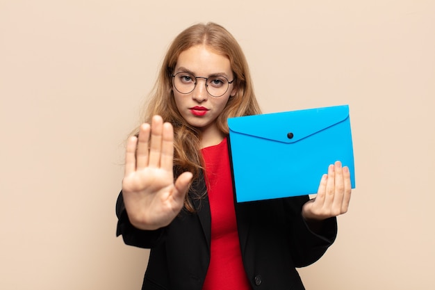 Blonde vrouw die ernstig, streng, ontevreden en boos kijkt die open palm toont die stopgebaar maakt