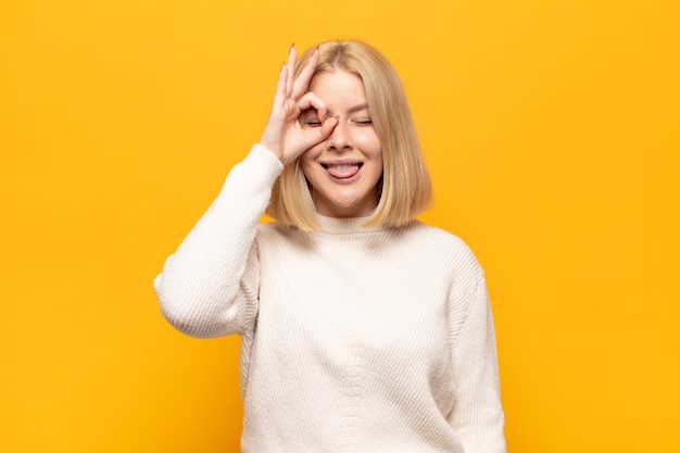 Blonde vrouw die gelukkig met grappig gezicht glimlacht, een grapje maakt en door kijkgaatje kijkt, geheimen bespioneert