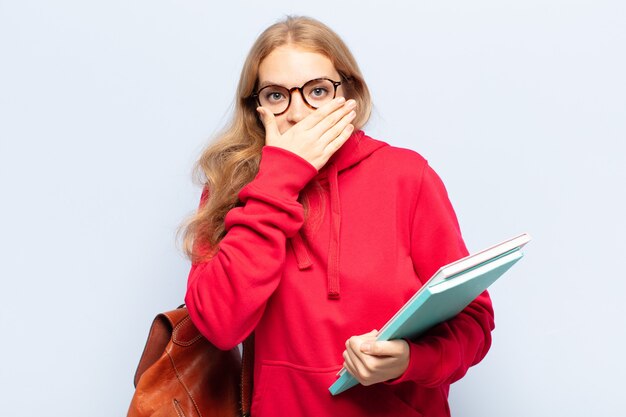 Blonde vrouw die mond bedekt met handen met een geschokte, verbaasde uitdrukking, een geheim bewaren of oeps zeggen