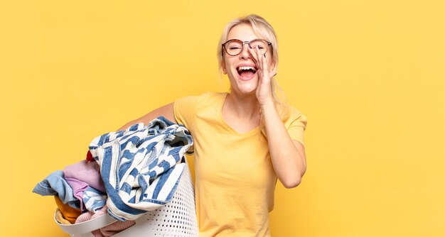 Blonde vrouw die zich gelukkig, opgewonden en positief voelt, een grote schreeuw geeft met de handen naast de mond, roept,