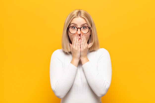 Blonde vrouw die zich ongerust, boos en bang voelt, de mond bedekt met handen, er angstig uitziet en het verprutst