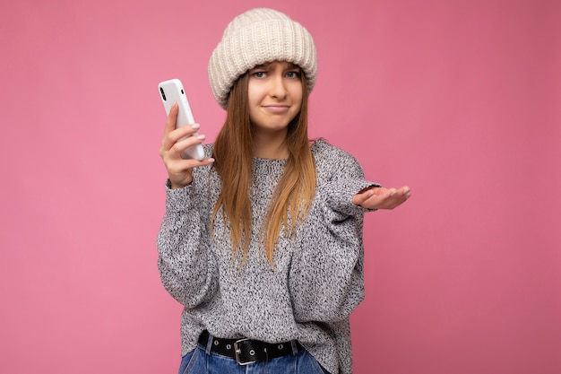 Blonde vrouw, gekleed in casual grijze trui en beige hoed geïsoleerd over roze muur in de hand houden en met behulp van mobiele telefoon en twijfels.