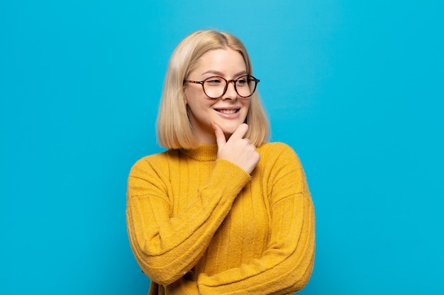 Blonde vrouw glimlachend met een gelukkige, zelfverzekerde uitdrukking met de hand op de kin, zich afvragend en opzij kijkend