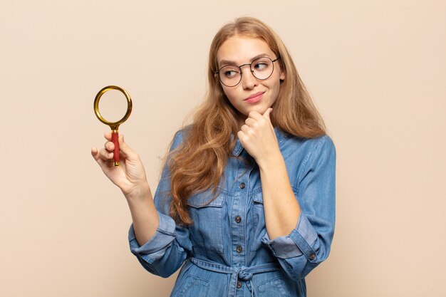 Blonde vrouw glimlachend met een gelukkige, zelfverzekerde uitdrukking met de hand op de kin, zich afvragend en opzij kijkend