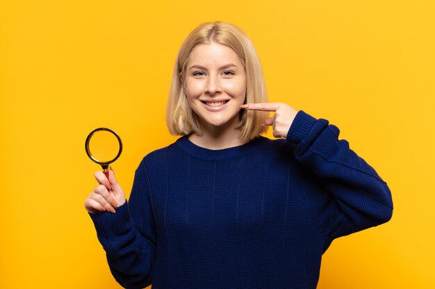 Blonde vrouw glimlachend vol vertrouwen wijzend op eigen brede glimlach, positieve, ontspannen, tevreden houding