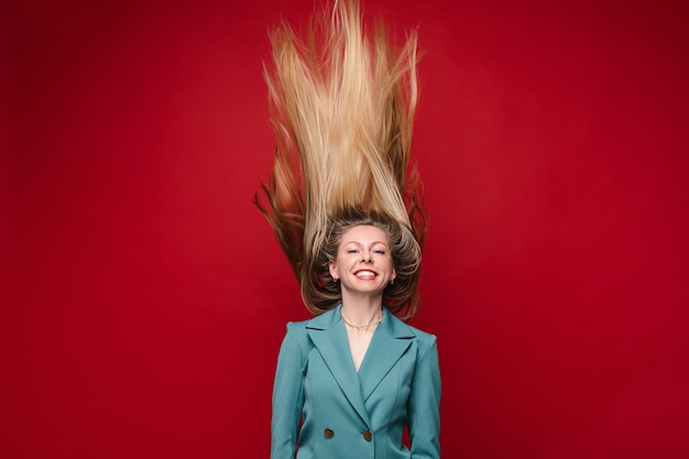 Blonde vrouw in blauwe jas plezier met haar haren in de lucht. Isoleer op rode achtergrond.