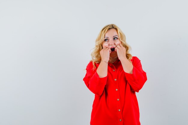 Blonde vrouw in rode blouse hand in hand in de buurt van mond, wegkijken en verbaasd kijken, vooraanzicht.