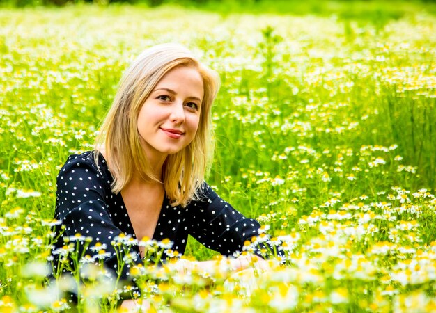 Blonde vrouw in zwarte jurk zit op chamomiles bloemen veld