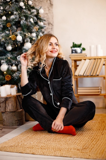 Blonde vrouw in zwarte pyjama zitten en poseren in de buurt van de kerstboom