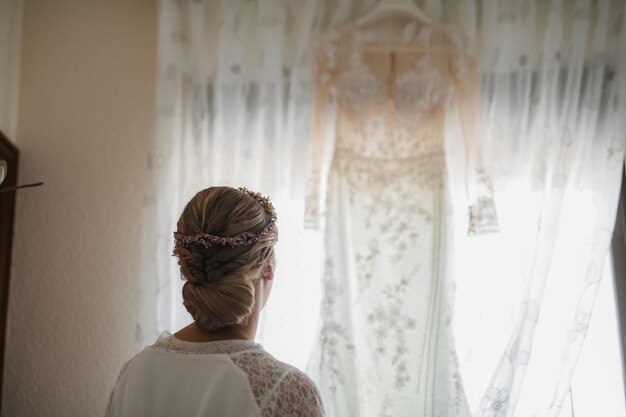 Foto blonde vrouw kijkt naar haar trouwjurk die in een raam hangt