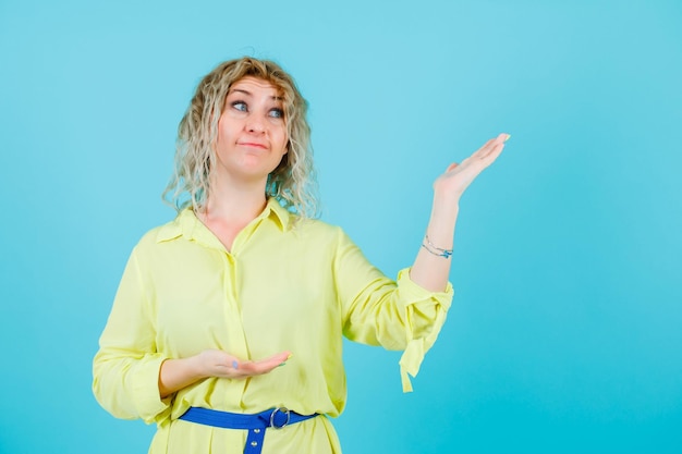 Blonde vrouw kijkt omhoog door haar hand op te steken op een blauwe achtergrond