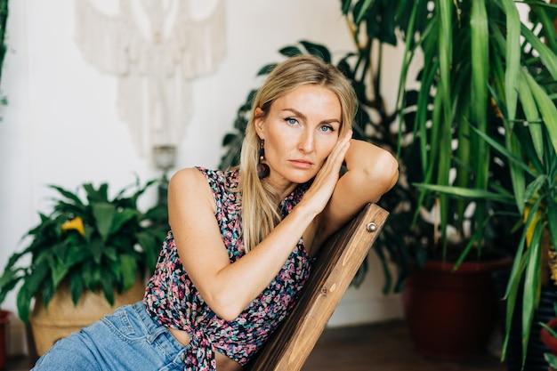 Blonde vrouw met blauwe ogen in een bloemenbloes die in een houten stoel zit en naar de camera kijkt