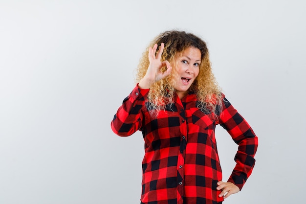 Blonde vrouw met krullend haar die een goed gebaar in een geruit overhemd toont en er tevreden uitziet, vooraanzicht.