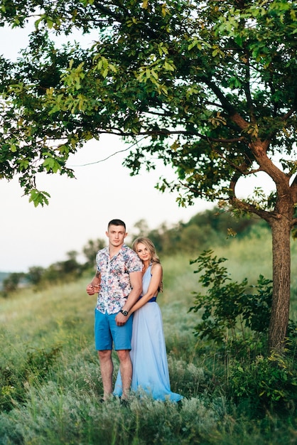 Blonde vrouw met los haar in een lichtblauwe jurk en een man in het licht van de zonsondergang in de natuur