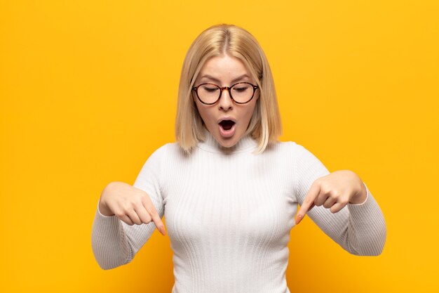 Blonde vrouw met open mond die met beide handen naar beneden wijst, geschokt, verbaasd en verrast