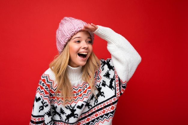 blonde vrouw met oprechte emoties die roze gebreide muts en wintertrui dragen die over rode achtergrond wordt geïsoleerd