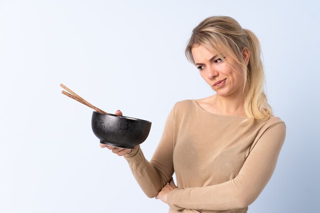 Blonde vrouw over geïsoleerd blauw met droevige uitdrukking terwijl ze een kom noedels met stokjes vasthoudt