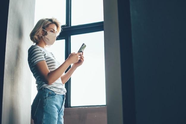 Blonde vrouw tijdens de lockdown zitten in de buurt van het raam en chatten op mobiele medische masker dragen